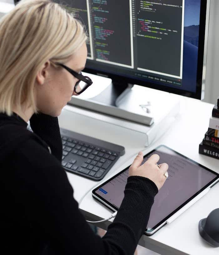 Woman in glasses working at a computer with code on screen and a tablet