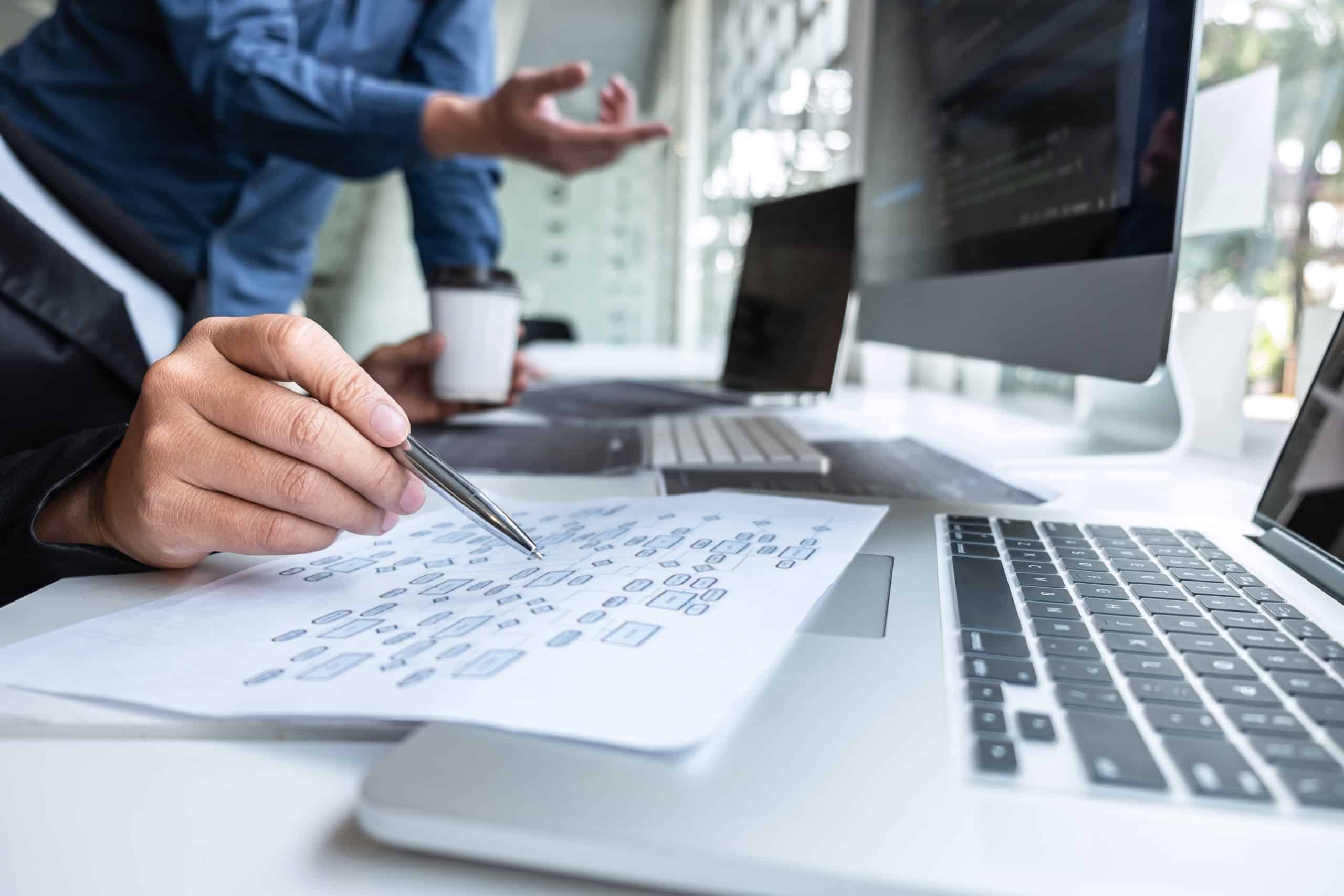 Hand pointing a pen at a process flowchart beside laptops and a monitor