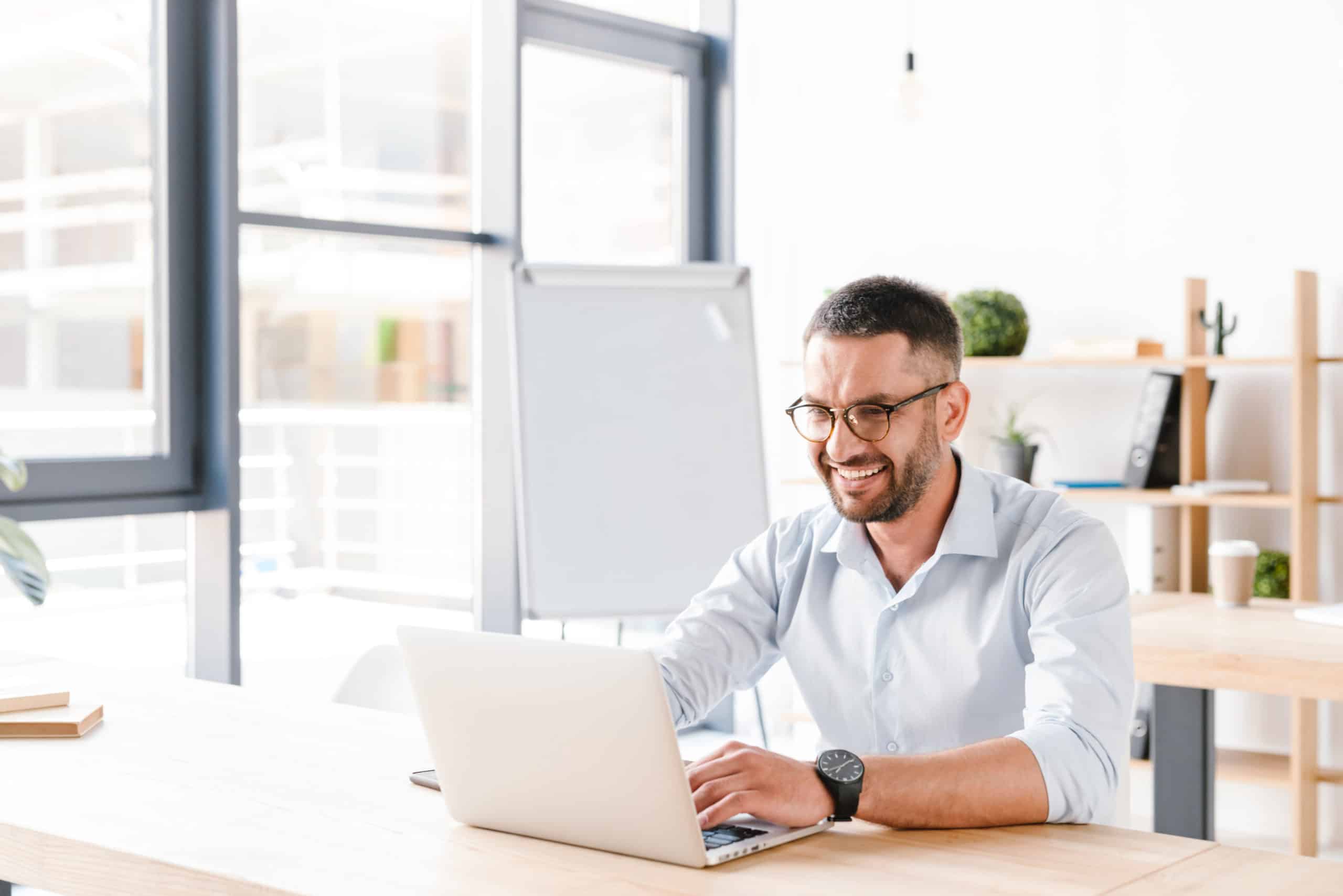Photo of joyous office man 30s in white shirt sitting at desk, and working on laptop